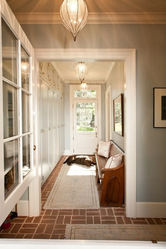 sleeping dog lying on beige rug, near a front door, inside hall with pale blue walls, and brown tiled floors, long hallway runners, various cupboards and a wooden settee