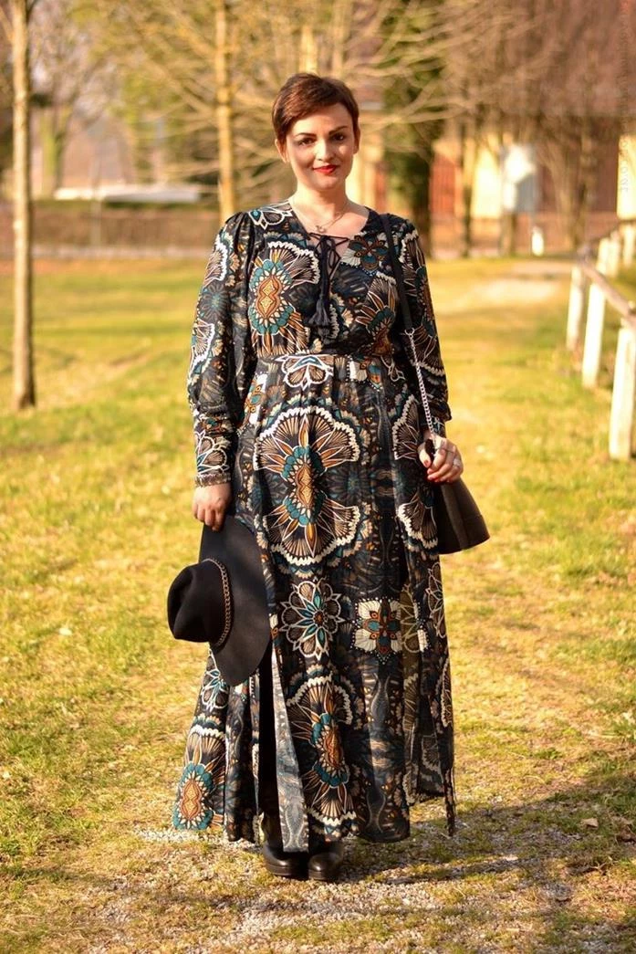 short-haired smiling woman, with pixie cut, wearing black maxi dress, with long sleeves, and colorful floral pattern, holding large bohemian style hat