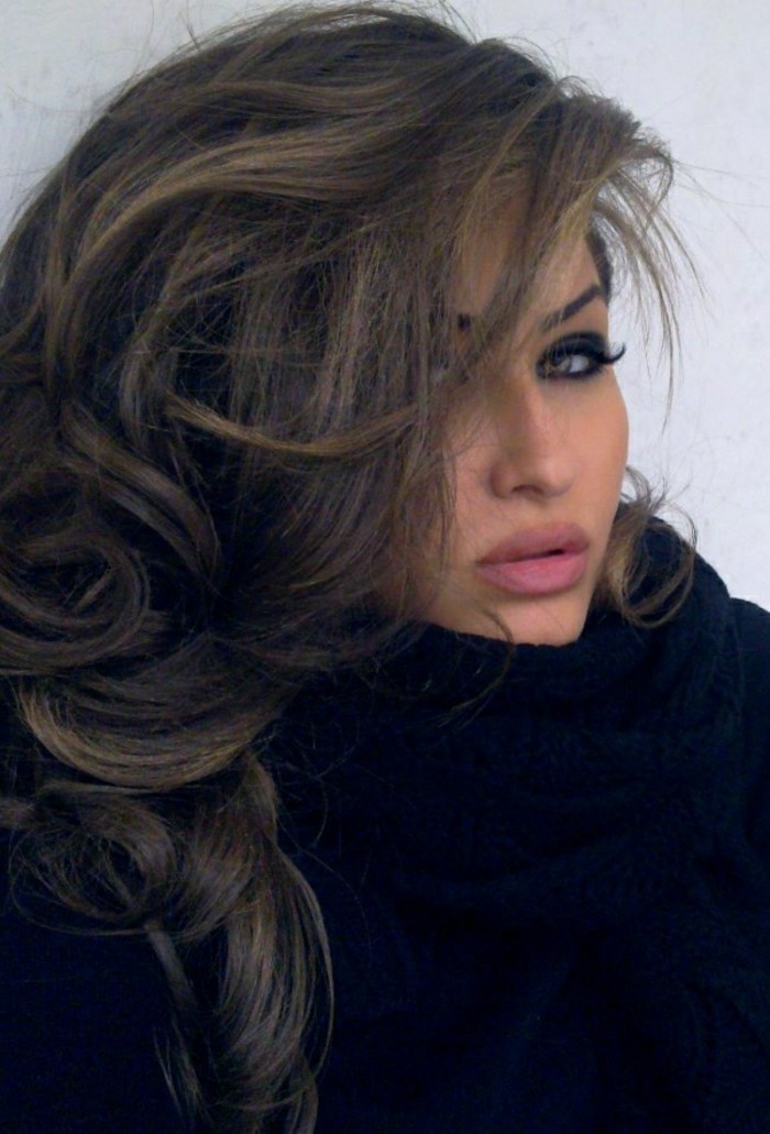 voluminous side swept and layered, brown highlighted hair, with dark blond strands, on woman with pink lipstick, and bold eye make up, with fake eyelashes, and chunky knit black scarf