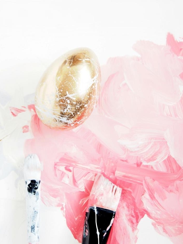 flat paintbrushes covered in pink and white paint, near a golden dyed egg, covered in white splashes of paint, easter egg decorating 