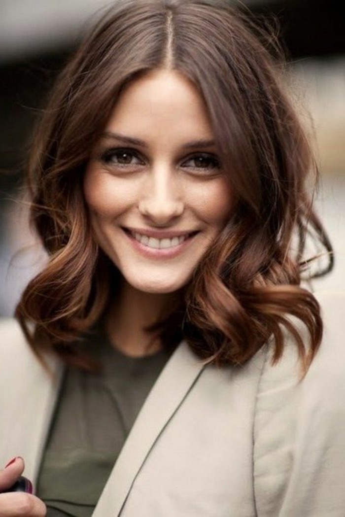 happy woman with curled, shoulder length hair, and medium brown hair color, middle parting, wearing discreet make up, beige blazer and a khaki top