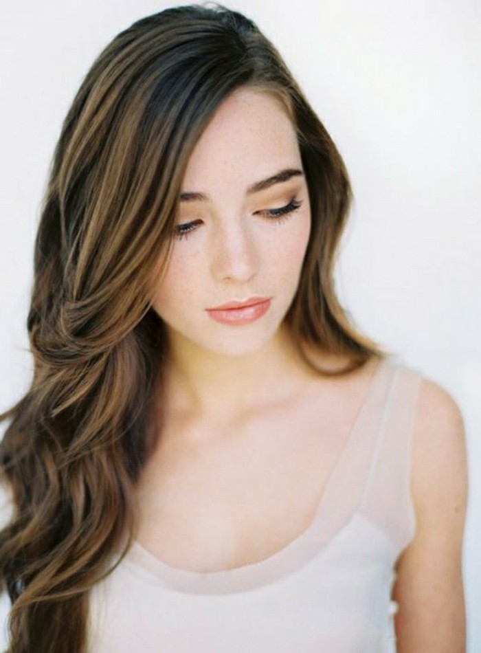thoughtful-looking young woman, with pink lipstick, and discreet eye make up, side-parted long and wavy, brown highlighted hair, and white tank top