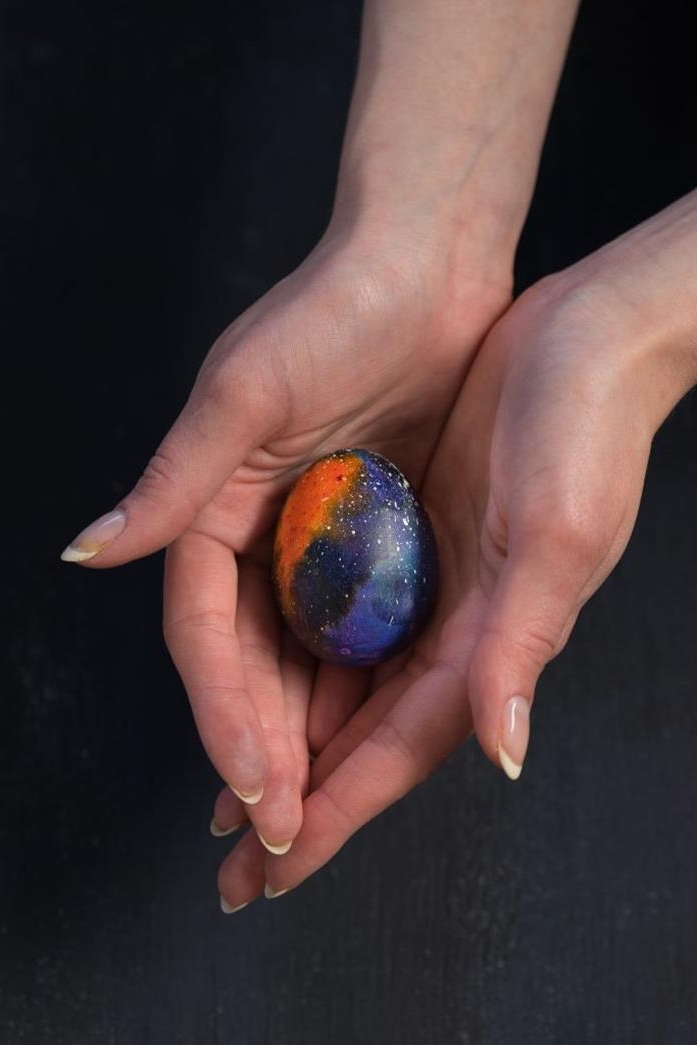 woman's hands with french manicure, holding a single easter egg, painted to look like deep space, how to dye easter eggs, on a black background