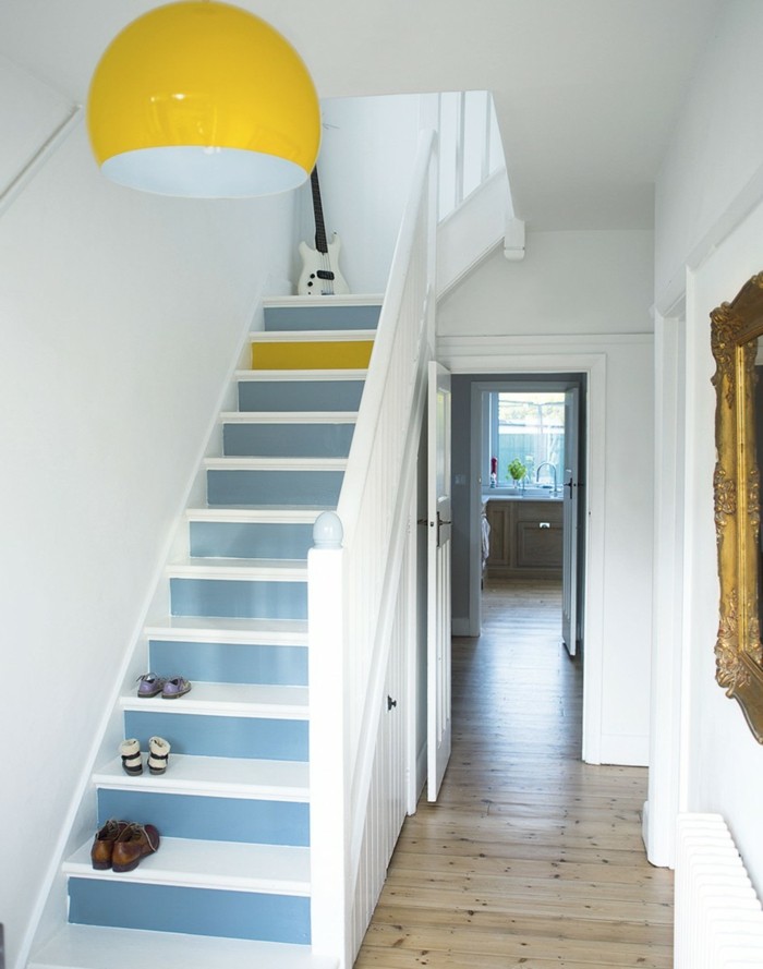 stairs painted in white and pastel blue, with a single step painted yellow, three pairs of shoes and a white electric guitar, large round yellow and white statement lamp, hallway decor, pale wooden floors, ornate golden frame 