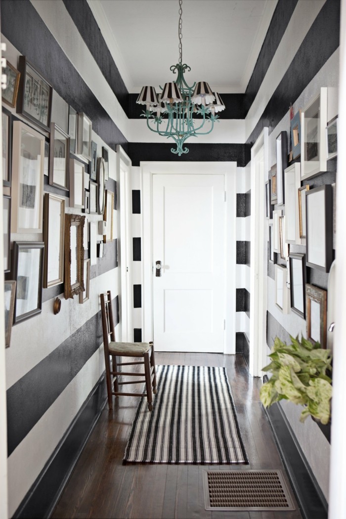 corridor painted in white and black stripes, with many images in different frames, small hallway ideas, dark wooden floor with striped rug, one wooden chair, blue chandelier with several striped lampshades