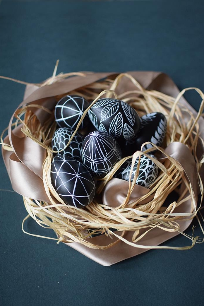 shiny black eggs, decorated with white drawings, of leaves and scales, geometric shapes and other patterns, dying easter eggs, placed in a nest made of straw, and a pale pastel pink ribbon