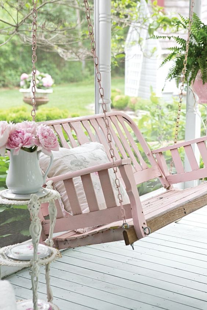 bench swing painted in pastel pink, hanging from pink chains, near a small white antique ornamental table, with a white jug, containing pink peonies