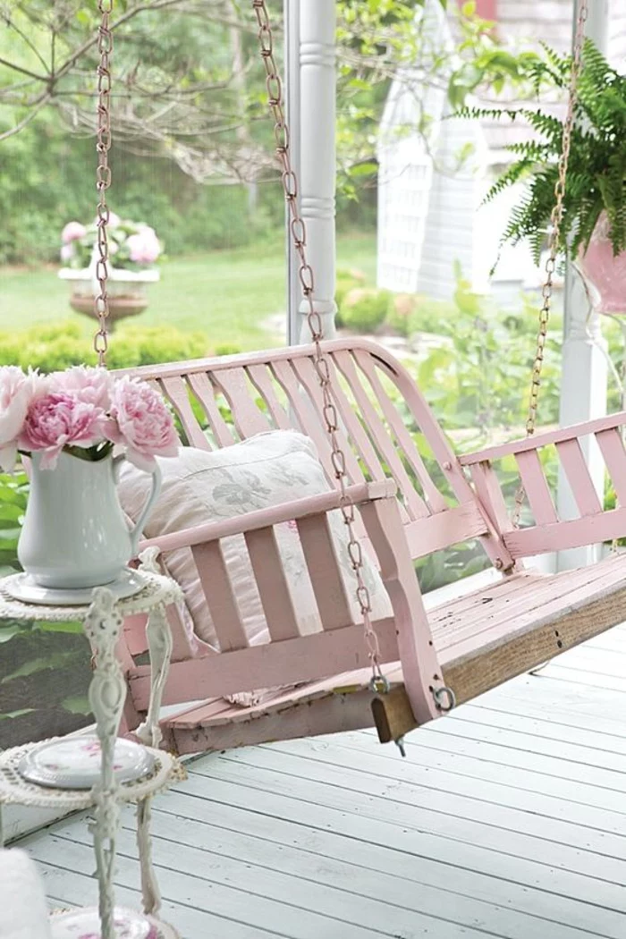 bench swing painted in pastel pink, hanging from pink chains, near a small white antique ornamental table, with a white jug, containing pink peonies