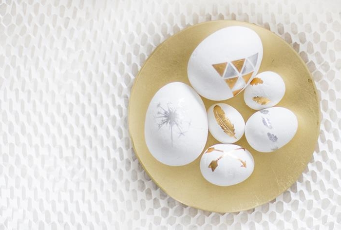 differently sized white eggs, covered with gold and silver tattoos, featuring geometric shapes, arrows and feathers, a sparkler and others, easter egg decorating, placed inside a round, flat golden dish