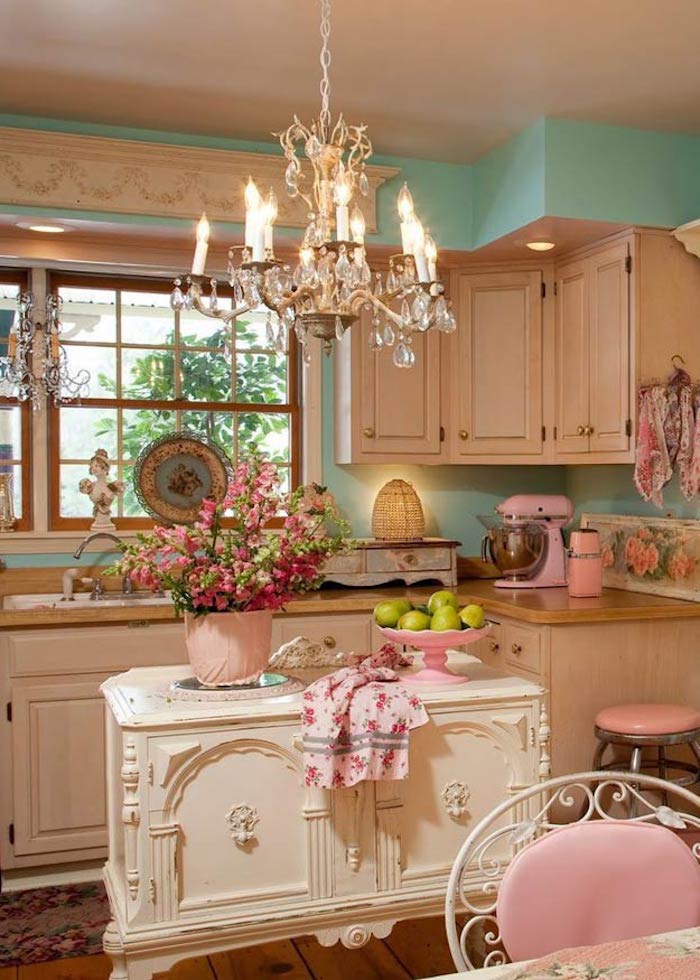crystal chandelier hanging in shabby chic kitchen, pastel blue walls and pastel pink furniture, white ornamental kitchen island, many decorative items and a flower pot