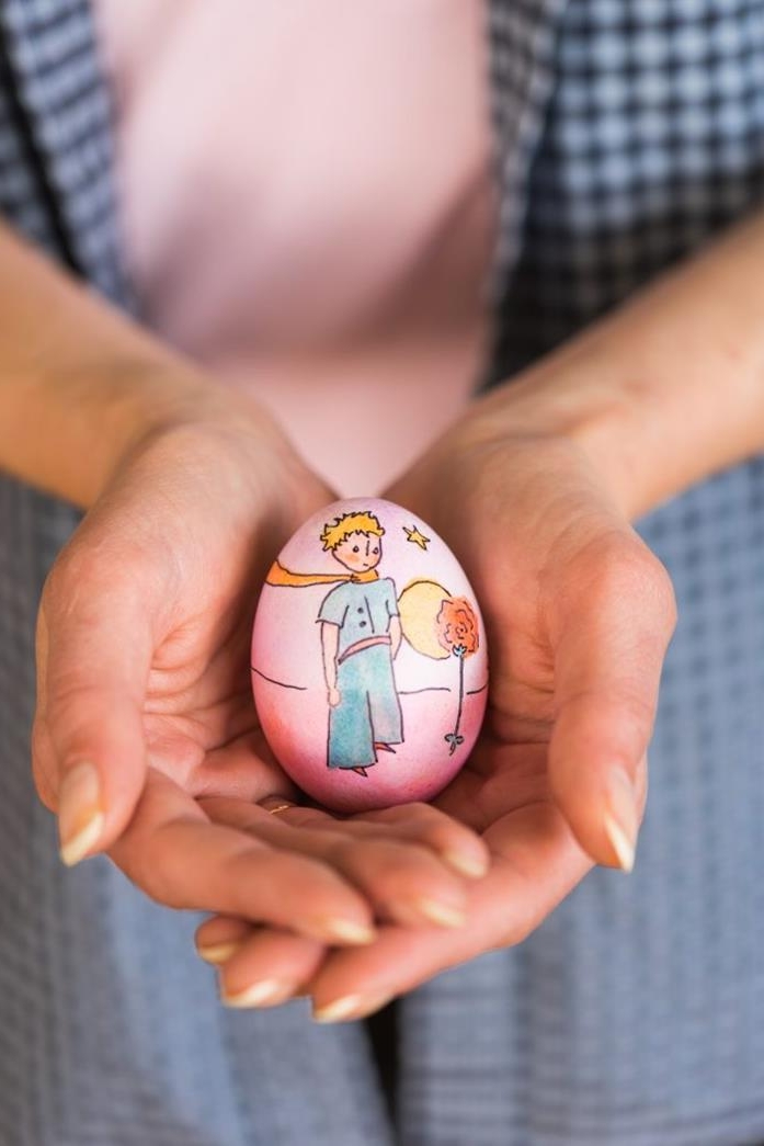 simple drawing of Antoine de Saint-Exupéry's the little prince, in full color, done on a pink easter egg, held by two hands