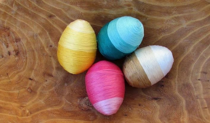 colorful thread in different shades of pink, yellow teal and brown, wrapped around four eggs, easter egg decorating, placed on wooden surface