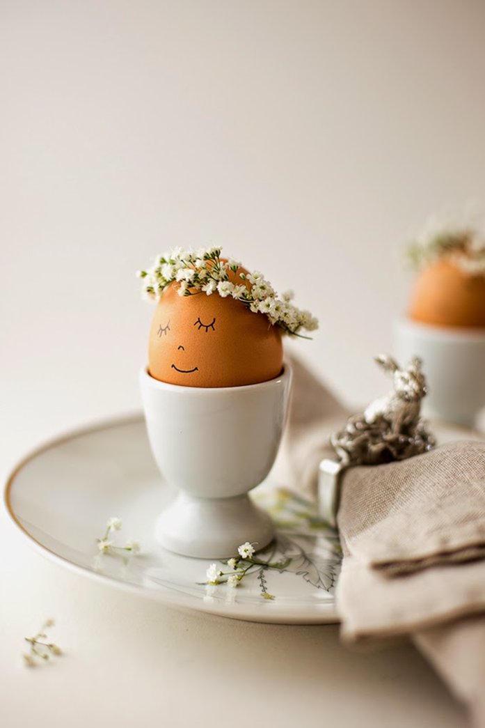 plain brown egg, with a tiny white flower crown, and minimalistic face, drawn on it in black sharpie, easter egg designs, placed in a white ceramic egg dish, on a plate