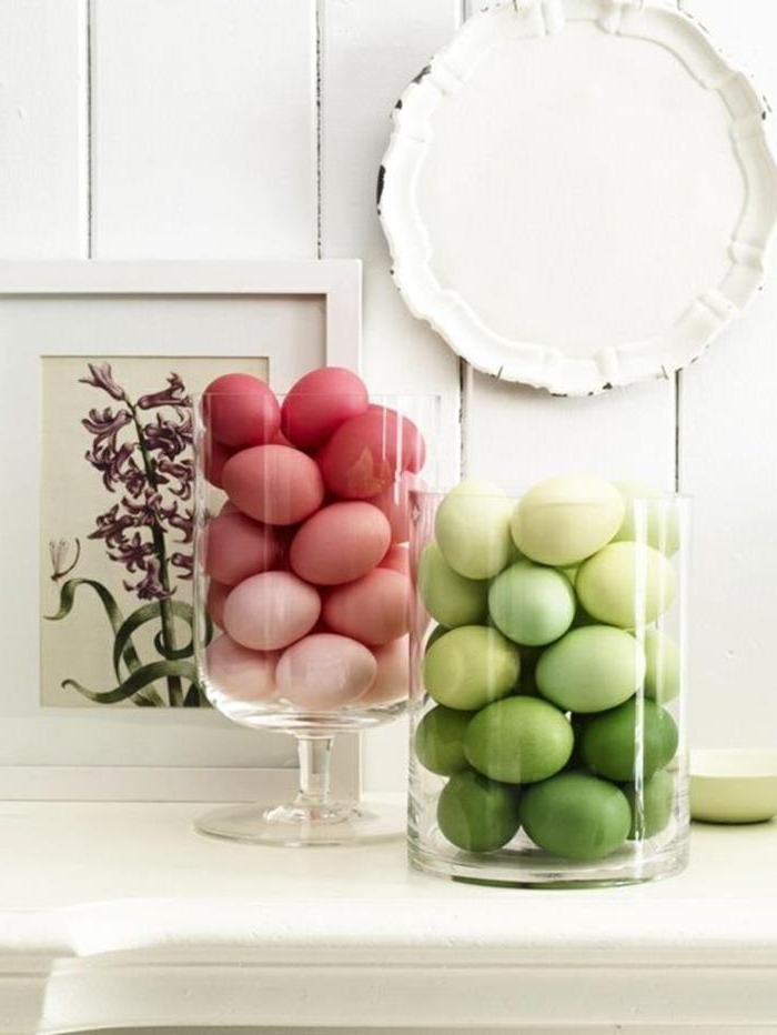 clear glass containers, filled with different shades of green, and red easter eggs respectively, placed on a white shelf, near a framed drawing of a flower, and a decorative plate