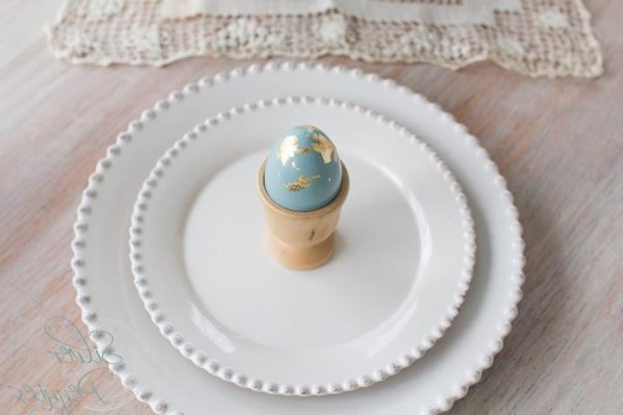 a single pale blue egg, decorated with gold leaf, placed in a beige egg dish, on top of two ornate, round white ceramic dishes