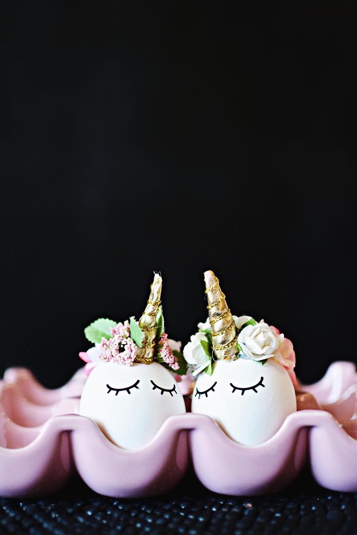 two white eggs, decorated to look like unicorns, with small golden fabric horns, little artificial flower crowns, closed eyes stickers, dying easter eggs, inside a pink egg dish