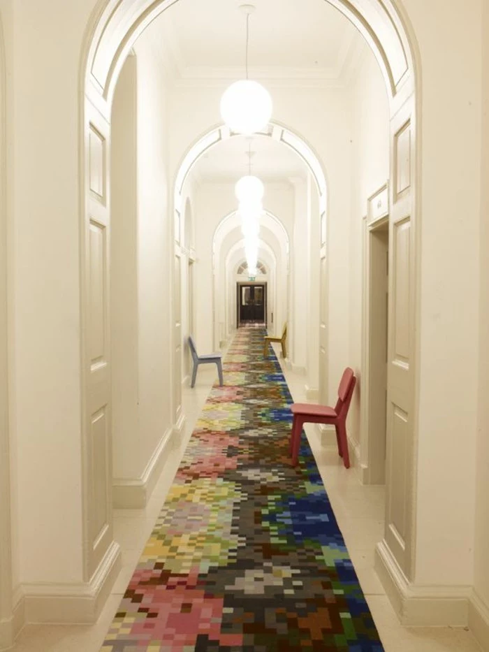 narrow and very long corridor, with white walls and several white doors, lit round ceiling lamps, three chairs in different colors, long hallway runners, multicolored rug covering the floor