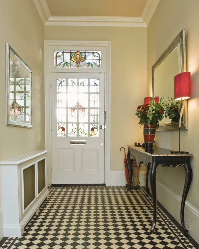 stained glass details on a white door, cream walls with white plaster details, black and white mosaic floor, hallway decorating ideas, black vintage table, with a flower vase and a lamp, near a mirror