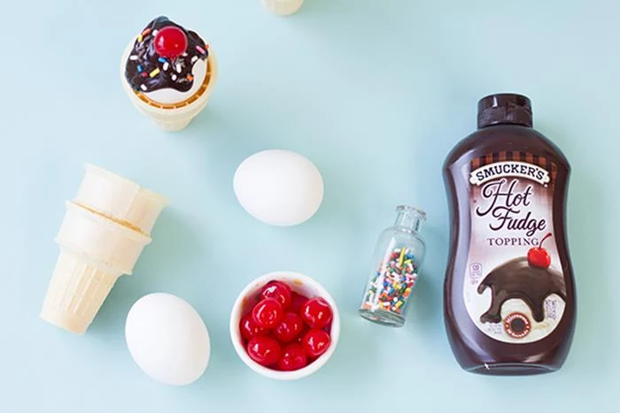 wafer ice cream cones, two white eggs, a small bowl of maraschino cherries, a little vial of sprinkles, a bottle of hot fudge topping, coloring easter eggs to look like ice-creams