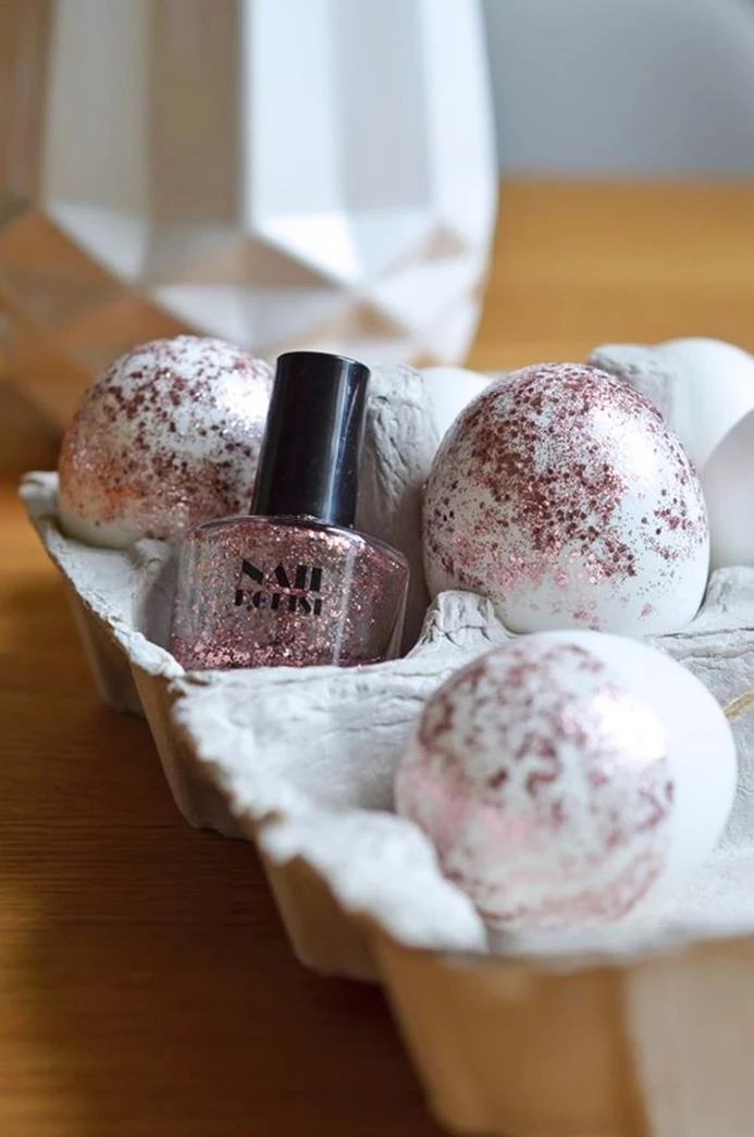 decorating eggs with nail polish, three white eggs, half-covered in clear nail polish, with pink glitter, placed in a cardboard egg box, near the nail polish bottle