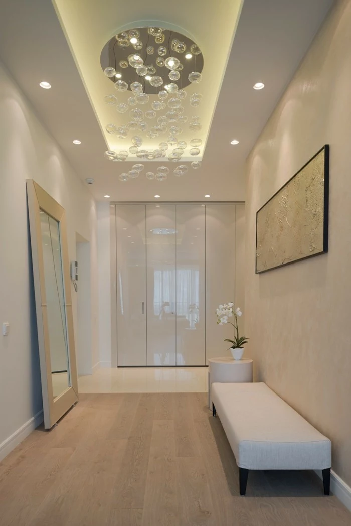 hallway decor, bright room with pale wooden floor, light cream walls, mirror in white frame, white settee and matching table, ceiling lamp with many crystal orbs, white framed painting
