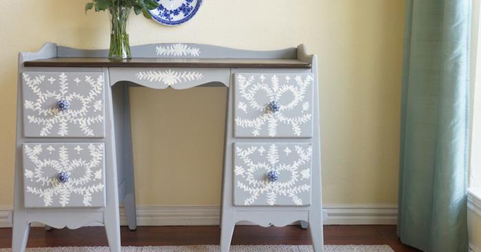 clear glass flower vase, on grey desk with drawers, hand-decorated with white patterns, near window with teal curtain