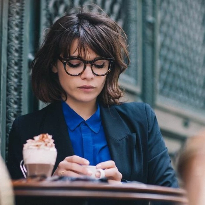 brunette hairstyles, woman wearing blue shirt, dark blue-green blazer, and glasses, with messy wavy bob and bangs, holding small white coffee cup