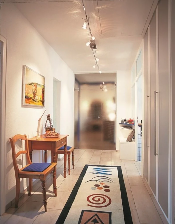 rug in beige and black, with several abstract shapes in different colors, long hallway runners, inside a hall with white walls, containing small wooden table and two chairs