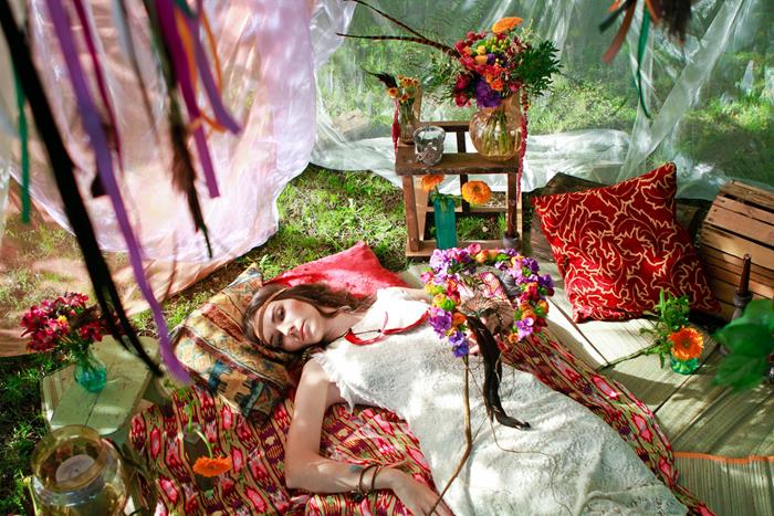 woman in long dress, made of white lace, lying on multicolored boho style, blanket, surrounded by cushions, flowers and ornaments