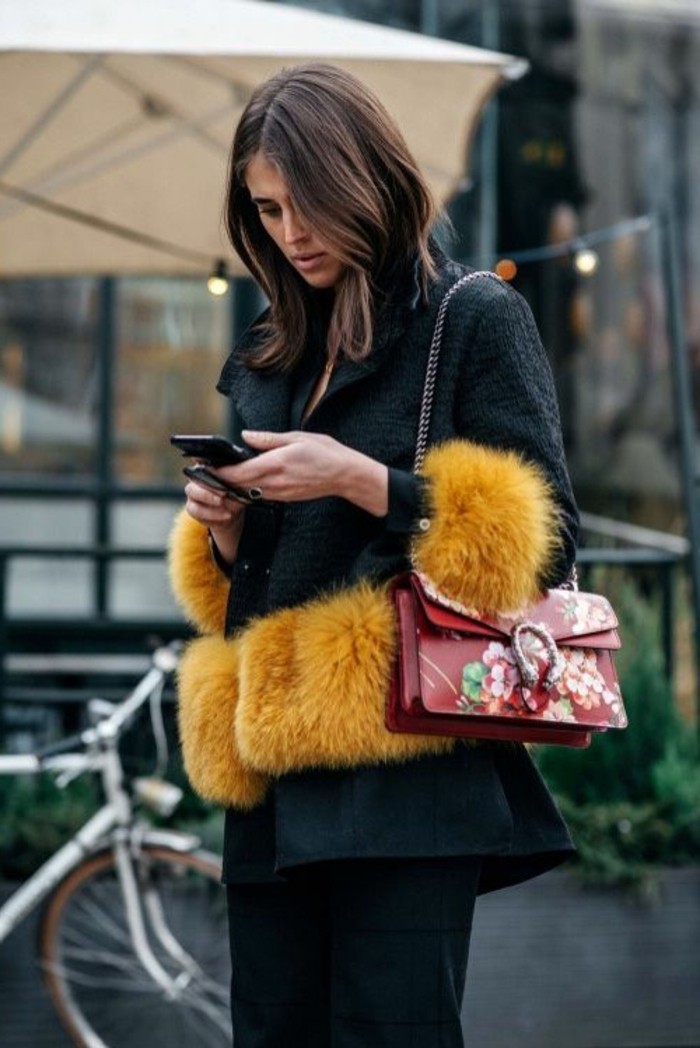 serious looking young woman, dressed in a dark green textured coat, with yellow fur trims, with dark trousers and a red bag, brunette hairstyles, shoulder length wavy hair