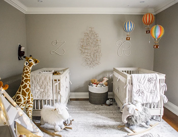 rocking gray elephant, and fluffy white horse, large stuffed giraffe toy, two white cribs, floating air balloon decorations, in room with gray walls, twin nursery ideas 