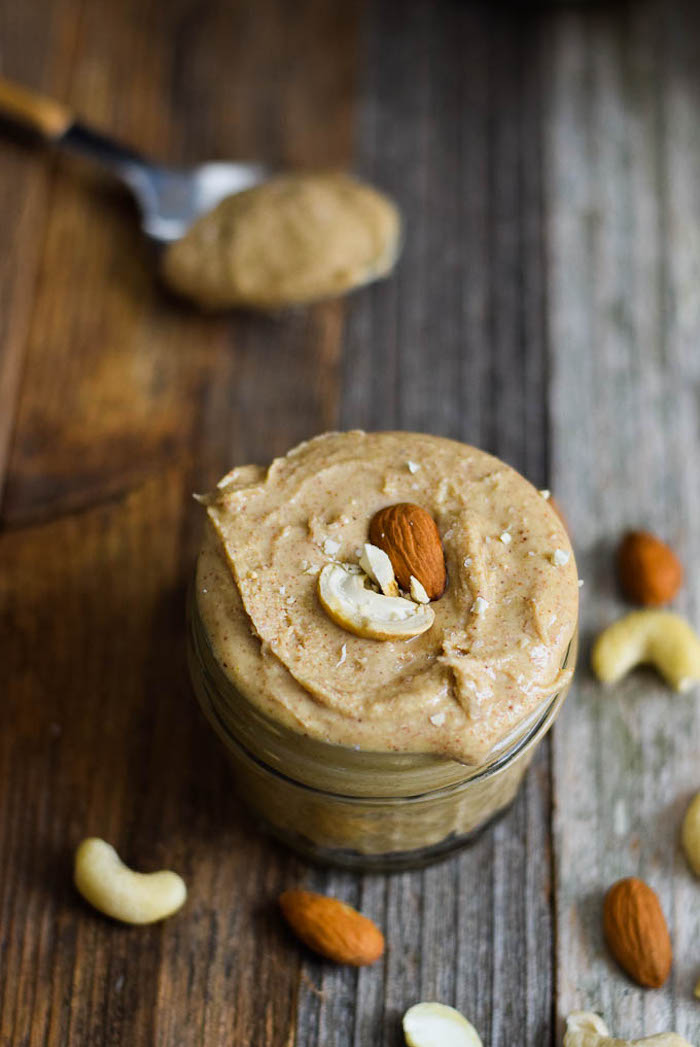 cashew and almond puree, topped with raw nuts, inside a small jar, on a wooden surface, with a spoon in the background