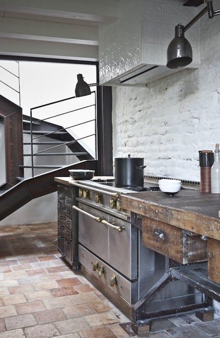 worn wooden cupboard, near black antique stove, with a vintage cooking pot, beige stone tiled floor, brick wall painted white