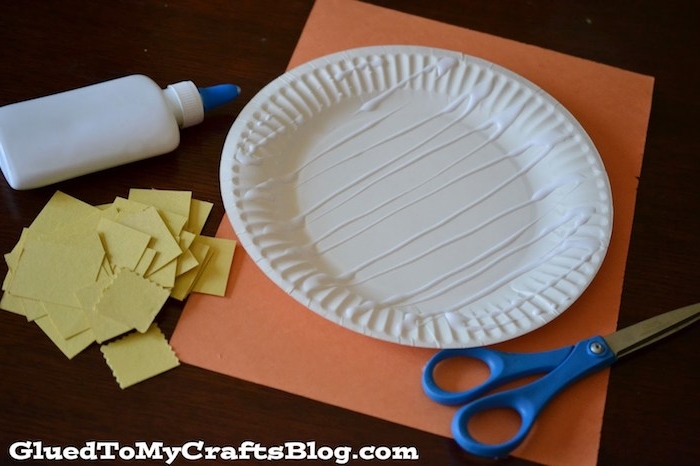 glue spread on round paper plate, resting on sheet of orange card, easter crafts for preschoolers, near blue scissors, glue bottle and many pieces of yellow paper