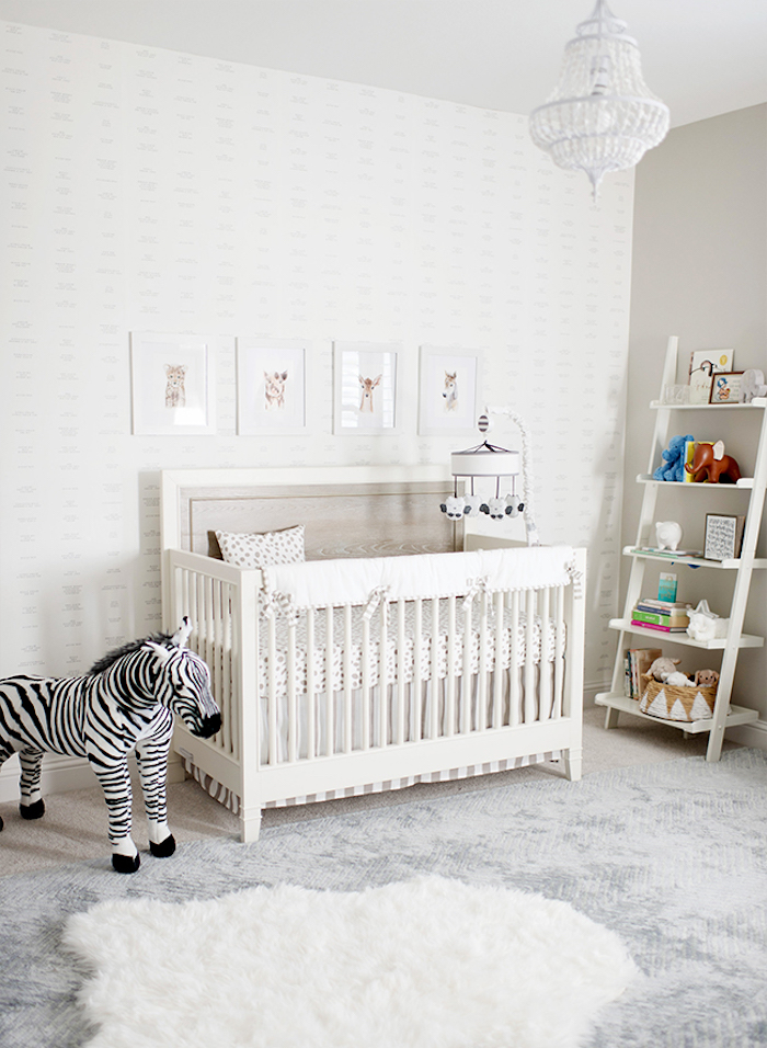 chandelier in white, inside baby room in light neutral colors, girl nursery themes, white crib near stuffed zebra toy, pale wallpaper and four framed drawings of animals