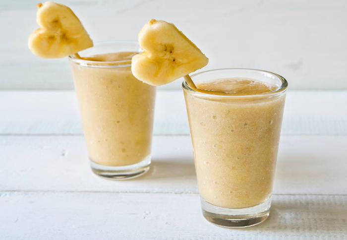 heart-shaped banana slices, on wooden sticks, decorating two glasses, filled with pale beige blended drink, fruit smoothie recipes, white wooden surface