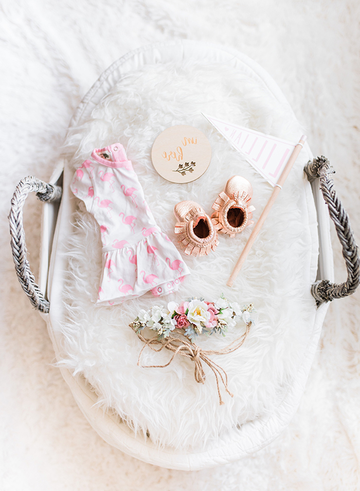 fluffy white carpet, baby girl nursery ideas, with pale gray basket, containing white plush cover, pale pink baby's dress, with flamingo pattern, tiny rose gold shoes, and others