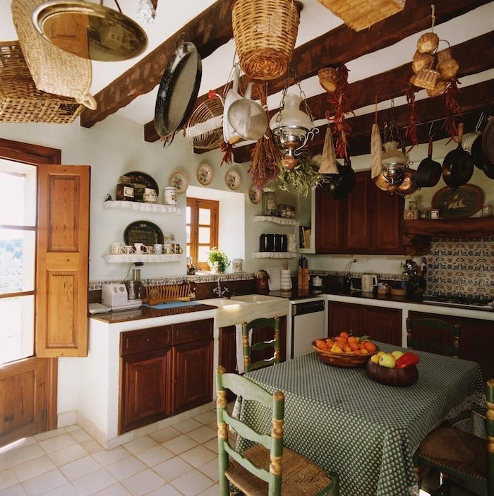 pans and baskets, and dried vegetables and herbs, hanging from the wooden beams of a ceiling, inside a room with rustic kitchen cabinets, small square table, with vintage green and beige chairs 