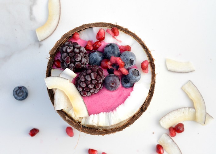 halved coconut filled with fruity, pale pink creamy substance, healthy breakfast smoothies, topped with blackberries, blueberries and pomegranate seeds, coconut chunks nearby