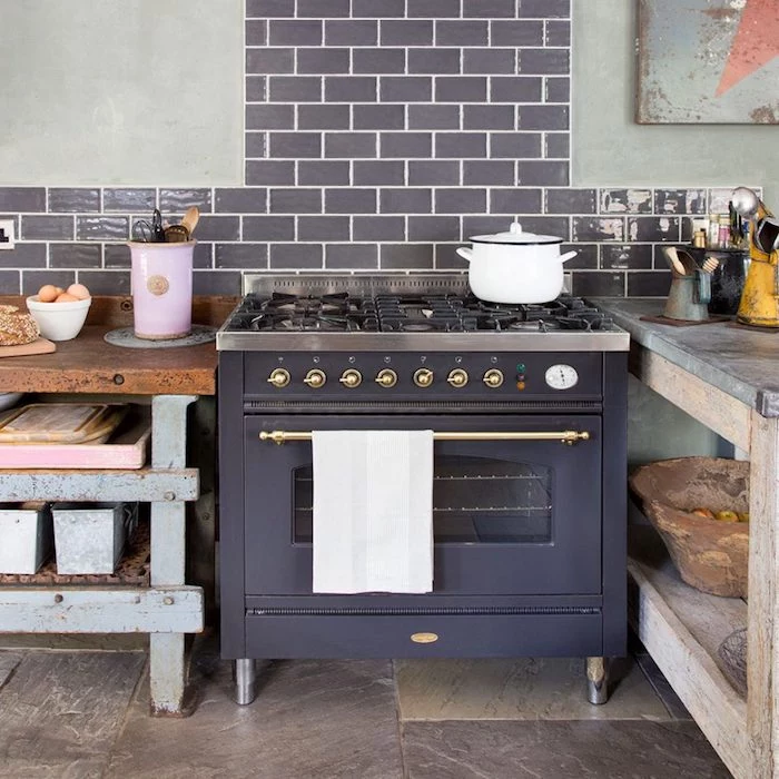 old fashioned black metal stove, in room with beige stone floor, pale gray walls, with dark gray brickwork details, shabby pale blue and brown, rustic kitchen cabinets 