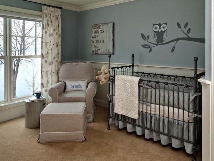metal ornamental crib in black, with gray and white bedding, near beige armchair, with matching foot stool, in baby nursery, with pale blue walls, white paneling and small dark gray mural