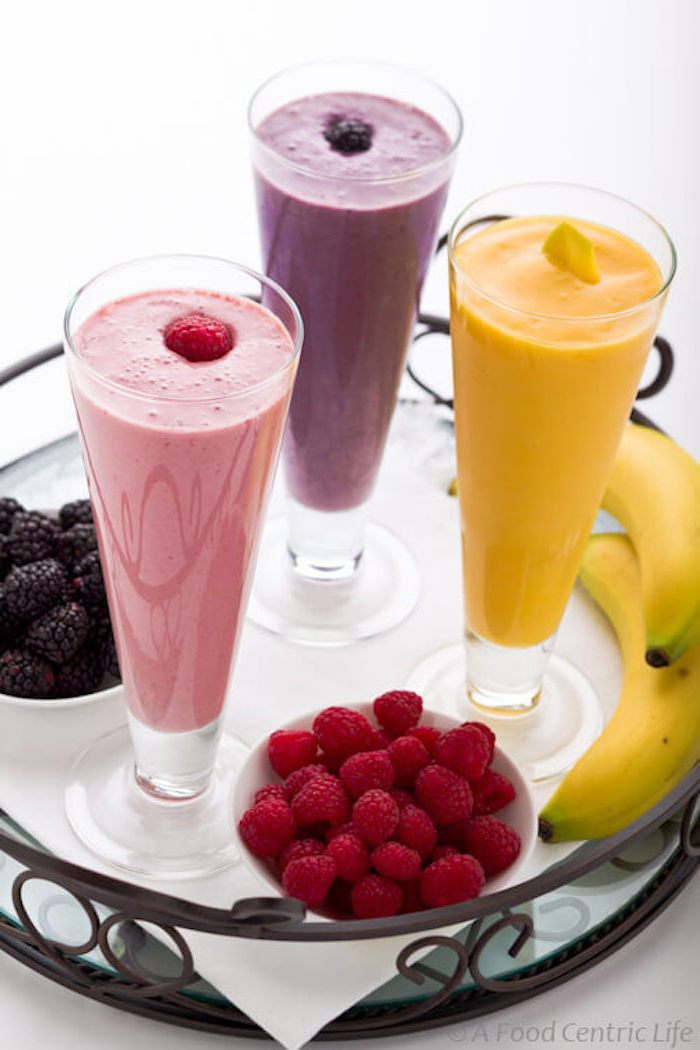 blackberries and strawberries in little white bowls, on tray containing two bananas, and three tall glasses, with blended drinks in different colors, smoothie recipes, each glass is topped with a different fruit