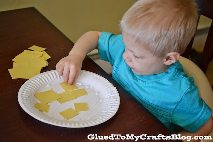 little blond boy, sticking yellow pieces of paper, on white paper plate, easter crafts for preschoolers, easy and fun idea