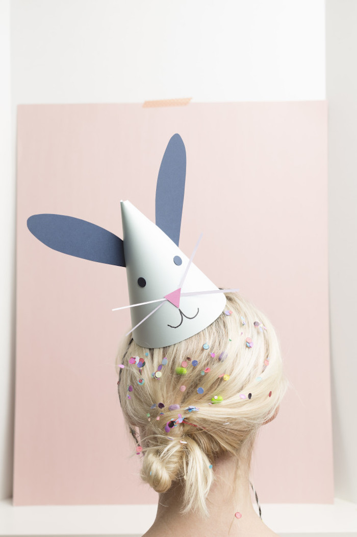 party paper hat, decorated with blue bunny ears, and a simple collaged face, on the head of a blonde woman, with tied hair, covered in colorful confetti and candy, cute crafts for adults and kids