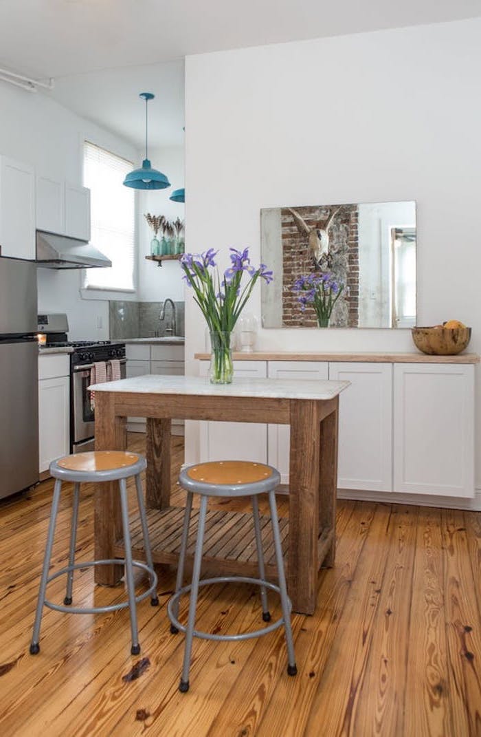 small wooden table, with flower vase, and two grey and brown stools, inside white kitchen, with light brown wooden floorboards, rustic home touches