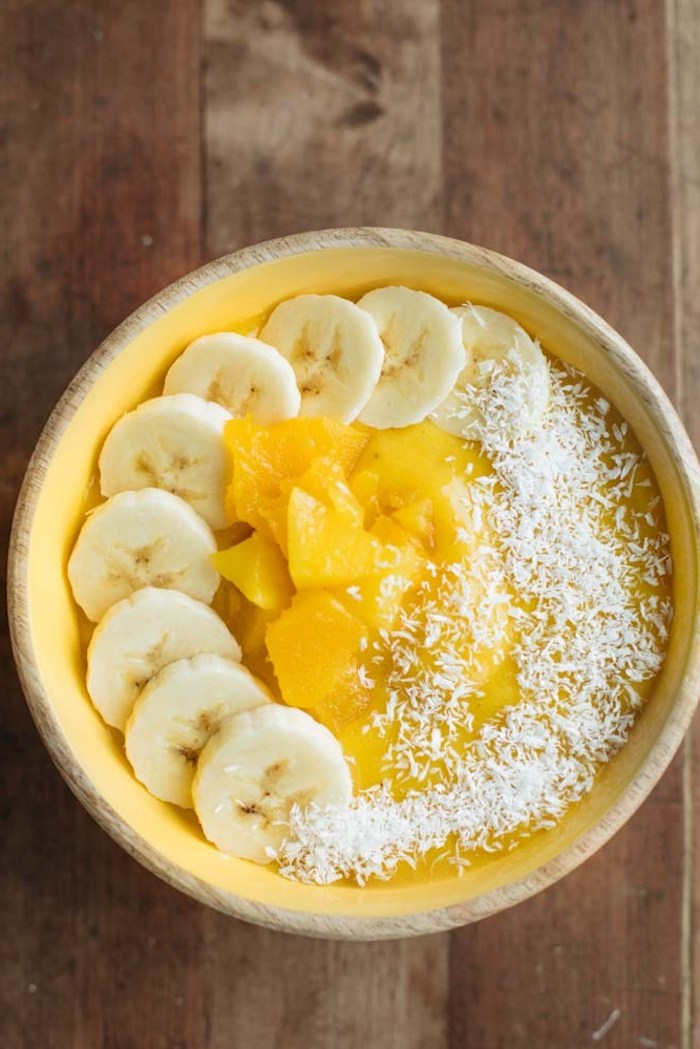 wooden bowl in yellow, containing yellow fruit puree, healthy breakfast smoothies, dusted with coconut shavings, and garnished with banana and mango slices
