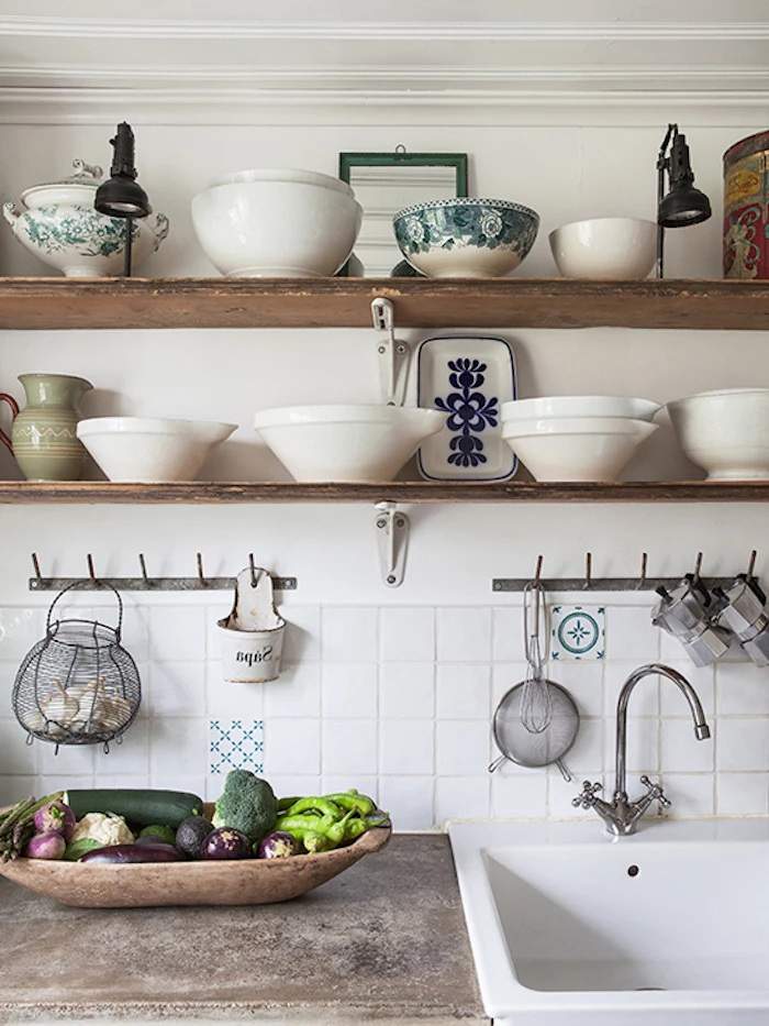 various white bowls, two lamps and a pale green pitcher, on two wooden shelves, above a vintage sink, inbuilt in a stone counter, vegetables in a large wooden dish