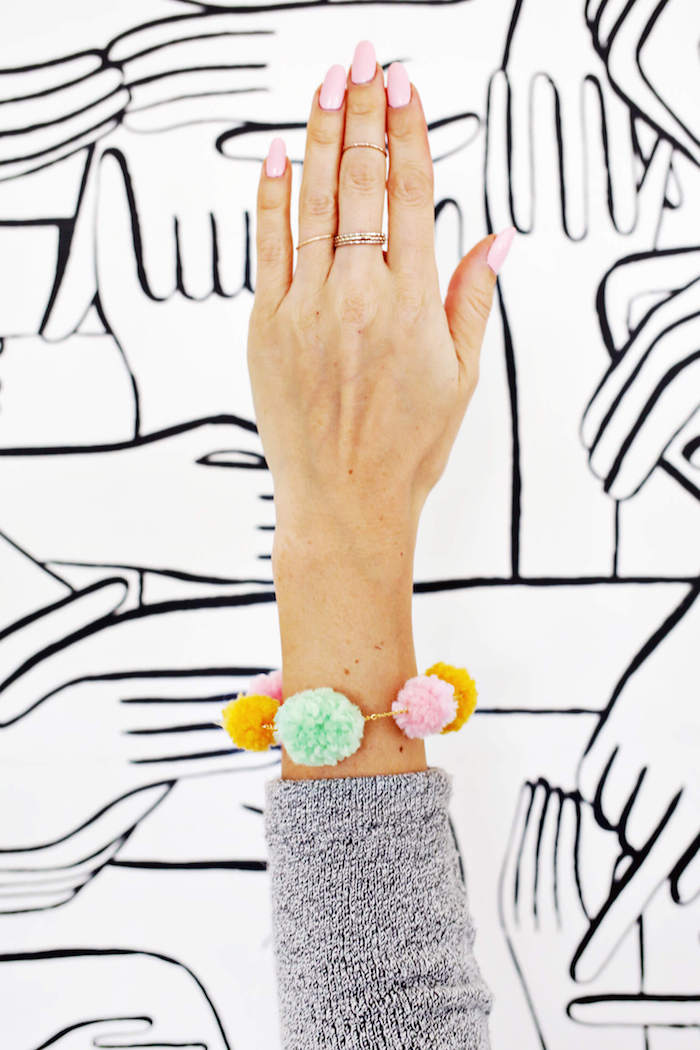 female arm with pale pink nail polish, and gray sleeve, wearing handmade pompom necklace, gift ideas for mom, in light green, pink and yellow