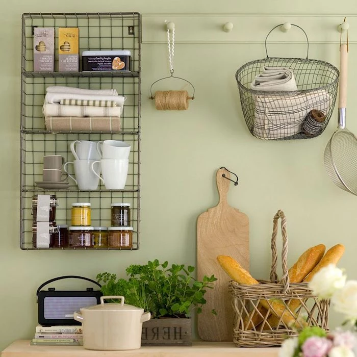 wire mesh shelves, covered with mugs, small jars and boxes of cookies, on pale pastel green wall, near counter with bread basket, planter with green herbs, and other rustic kitchen items
