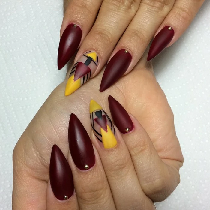 abstract symmetrical drawing, done in yellow, black and dark red, on dark wine-red manicure, long and sharp nails, tiny gold pearl details